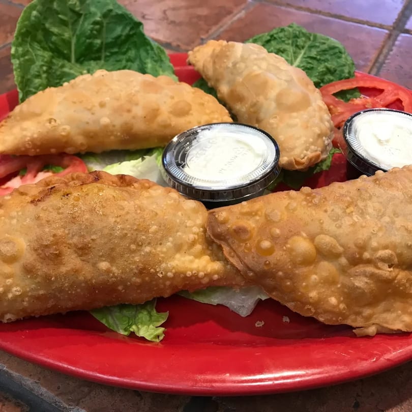 Four golden-brown fried empanadas on a red plate with lettuce, tomato, and two dipping sauces.