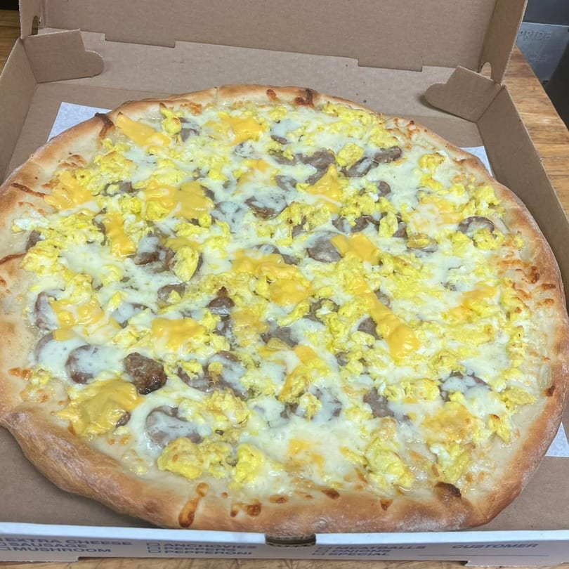 A breakfast pizza from Tj Pizza with scrambled eggs, sausage, and melted cheese in a box.