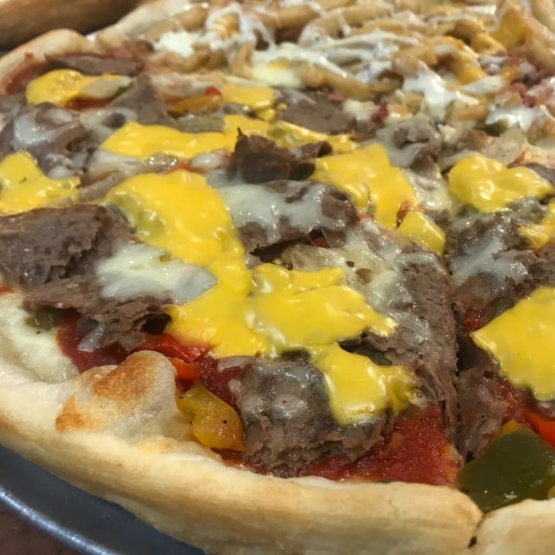 Close-up of a Tj Pizza cheesesteak pizza with steak, melted cheese, peppers, and onions.