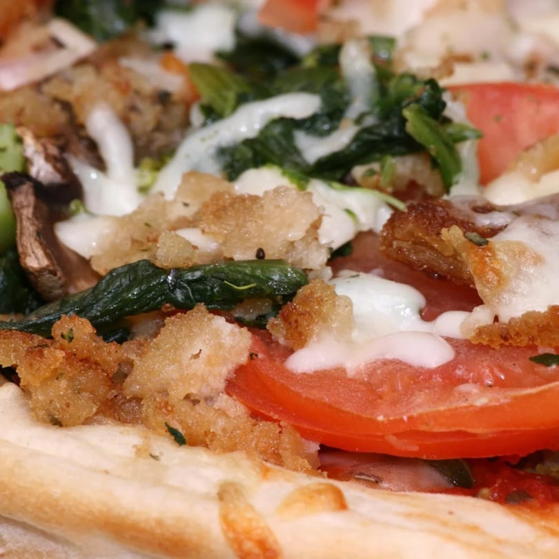 Close-up of a Tj Pizza slice with tomatoes, spinach, mushrooms, croutons, and white sauce.