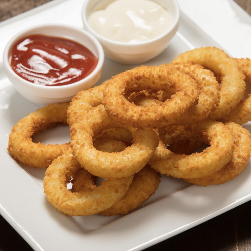Golden-brown onion rings on a white plate with small bowls of ketchup and mayonnaise.